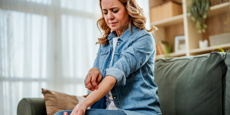 Woman sitting on a couch scratching her itchy forearm, wincing in discomfort